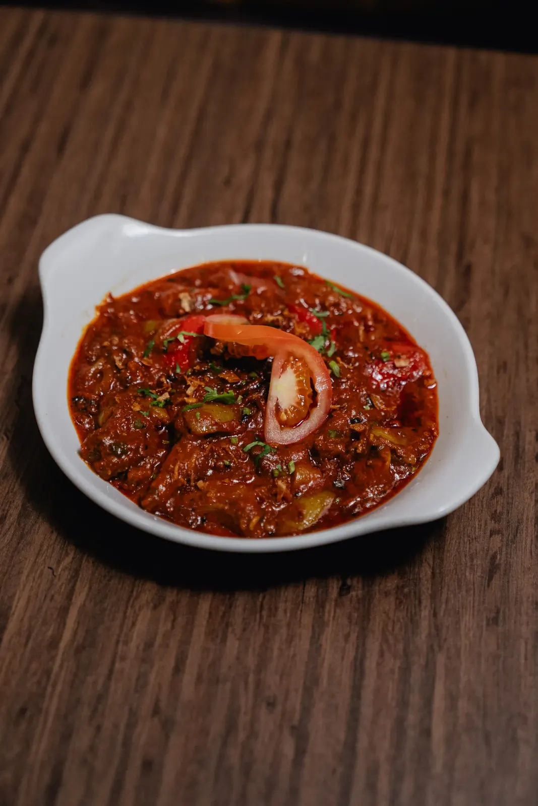 A rich tomato and pepper curry garnished with fresh tomato and coriander in a white serving dish