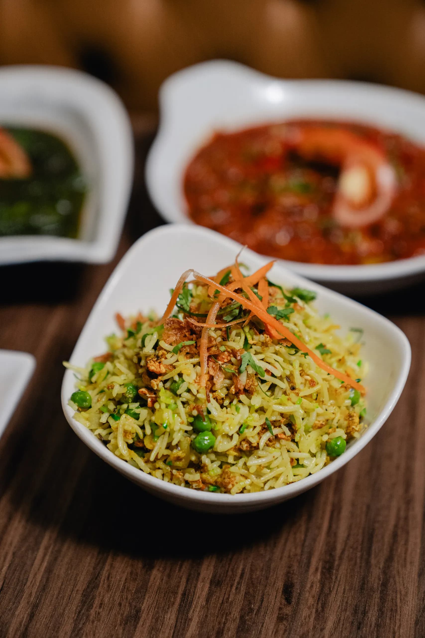 A bowl of vegetable pilau rice with peas and crispy fried onions, with a tomato curry and spinach saag behind
