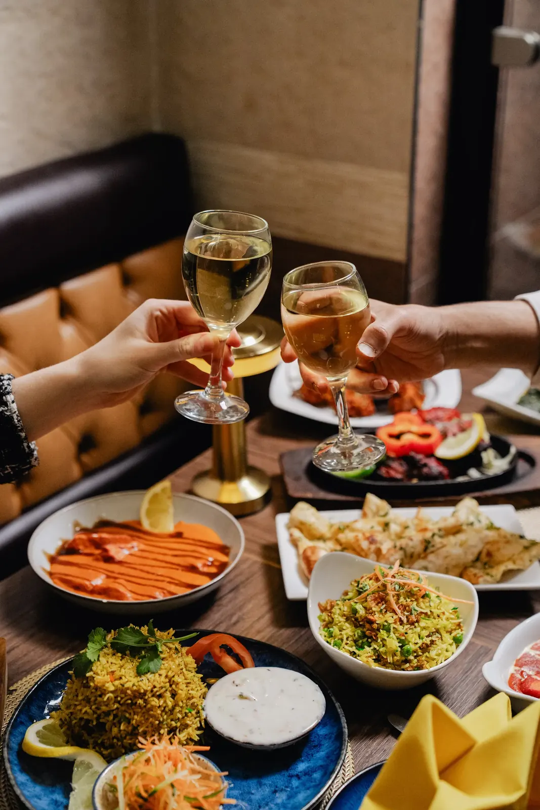 Two diners raising glasses of white wine over a spread of curries, pilau rice, raita and naan